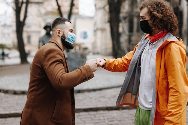 Dois jovens com máscaras protetoras médicas, cumprimentando com colisão de punho ao ar livre. Conceito de medida de quarentena e distância social.