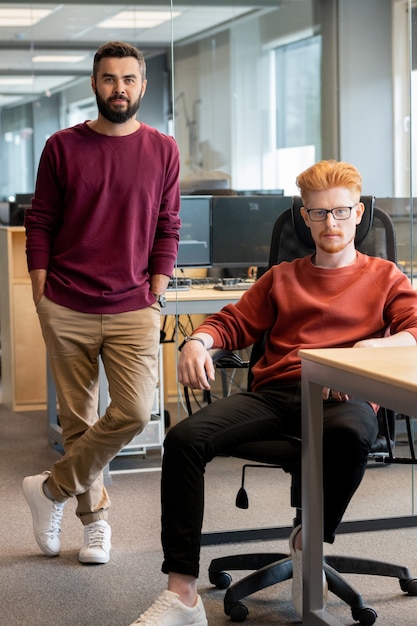 Dois jovens empresários em trajes casuais sentados e de pé perto do local de trabalho, contra monitores em um grande escritório contemporâneo