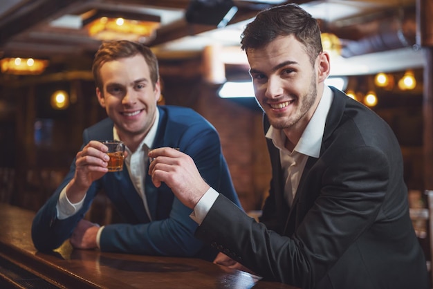 Dois jovens empresários estão sorrindo olhando para a câmera e batendo copos de bebida alcoólica juntos enquanto estão sentados no balcão de bar no pub