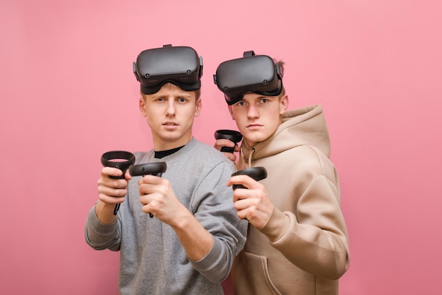 Dois jovens estão em um fundo rosa com capacetes VR em suas cabeças e controladores em suas mãos olhando atentamente para a câmera em um fundo rosa Amigos jogam jogos VR no console