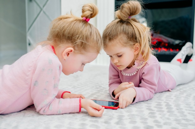 Dois jovens focados crianças jogando um smartphone sem nome deitado no chão da sala. Crianças pequenas e tecnologia, irmãs brincam com um telefone celular, assistem a vídeos ou jogam jogos