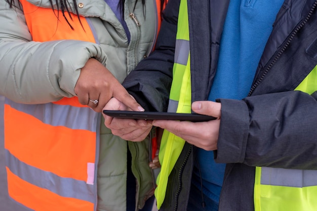 Dois jovens trabalhadores em coletes laranja e amarelos olhando para o tablet e discutem um projeto