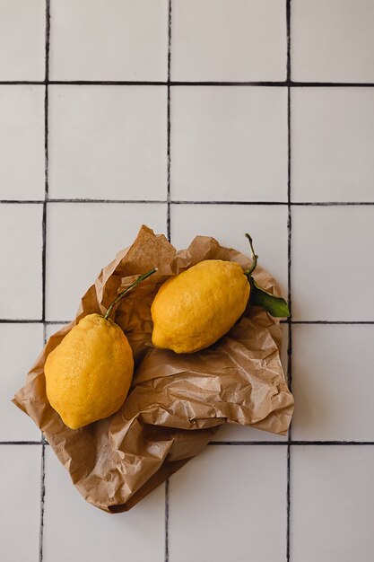 Dois limões grandes com uma folha verde em uma sacola de compras marrom rasgada em uma bancada de cozinha de azulejos brancos.