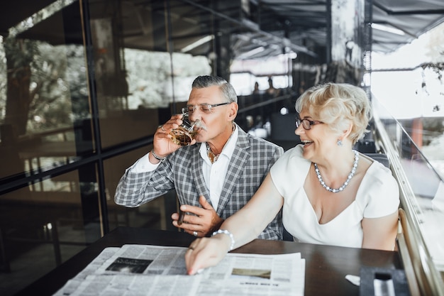 Dois lindos casais de idosos bebendo vinho no terraço de verão e lendo jornal neste momento