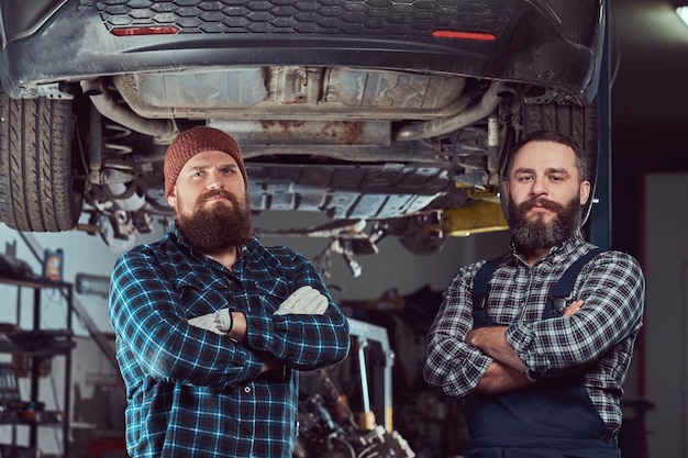 Dois mecânicos brutais barbudos em pé com os braços cruzados contra um carro em um elevador na garagem.