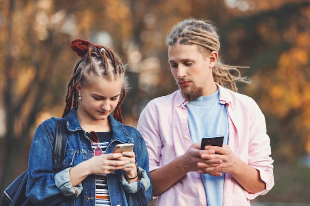 Dois melhores amigos hipster com dreads vestindo roupas casuais digitando mensagens em seus smartphones homem loiro barbudo olhando no smartphone de sua namorada vista ao ar livre