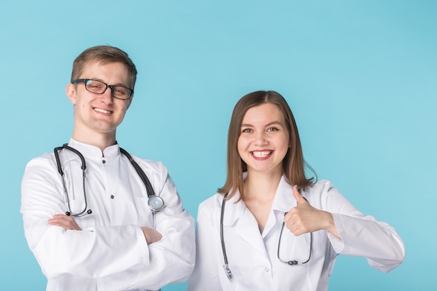 Dois melhores profissionais inteligentes sorrindo trabalhadores médicos em jalecos brancos, mostrando os polegares em pé contra a superfície azul.