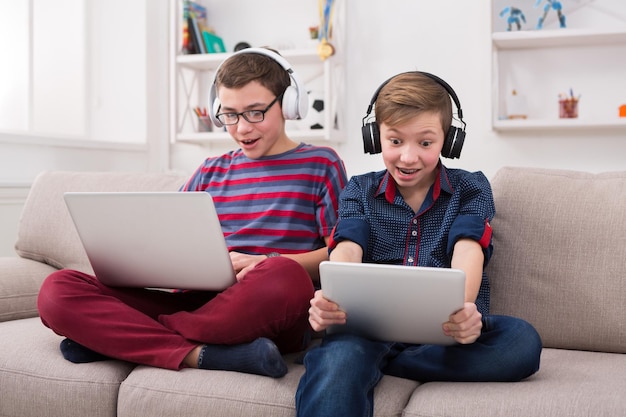 Dois meninos com aparelhos e fones de ouvido. Amigos adolescentes jogando no laptop e tablet digital com emoção em casa. Amizade de menino e conceito de jogo online