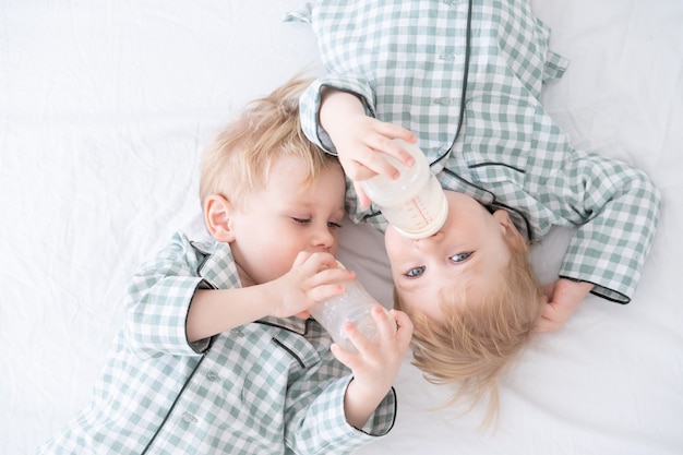 Dois meninos gêmeos do bebê da criança de pijama deitam na cama bebendo leite em garrafas. Vida familiar sincera de infância.