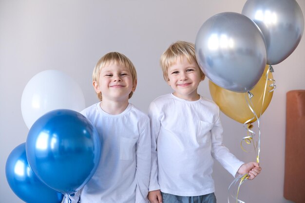 Dois meninos pequenos loiros sorridentes gêmeos abraçados segurando balões comemorando aniversário