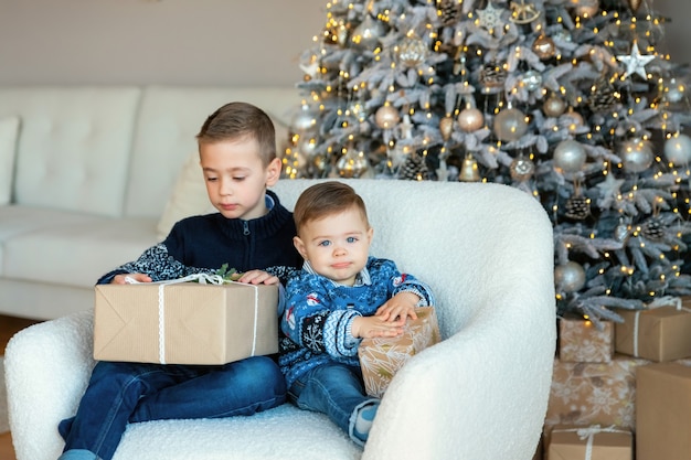 Dois meninos sentados em uma cadeira confortável com presentes nas mãos. em um fundo, a árvore do ano novo com ornamentos rústicos. manhã de natal na luminosa sala de estar.