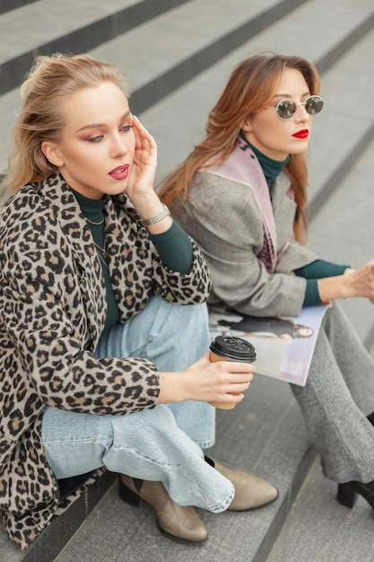 Dois modelos elegantemente bonitos de garotas hipster em roupas da moda sentam-se nos degraus da cidade