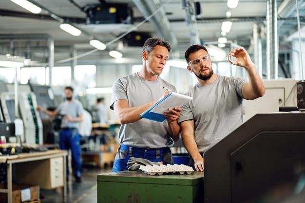 DOIS operadores de máquinas CNC examinando produtos acabados e escrevendo notas em instalações industriais