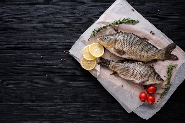 Dois peixes carpas estão deitados no papel em um fundo preto de madeira de tábuas, ao lado de verdes, tomates de alecrim e limão