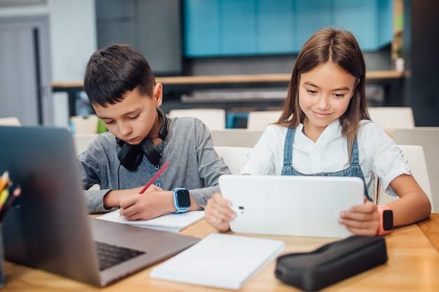 Dois pequenos amigos escrevendo dever de casa, usando o tablet digital inteligente na moderna sala de aula azul.