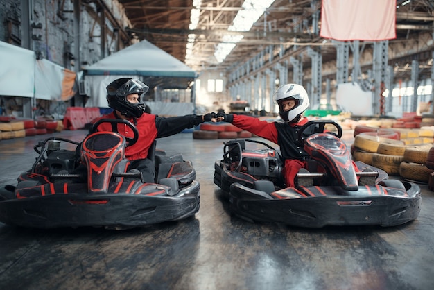 Foto dois pilotos de kart na linha de largada, vista frontal, karting automotivo interno. corrida de velocidade em pista fechada de kart com barreira de pneus. competição de veículos velozes, lazer com alta adrenalina