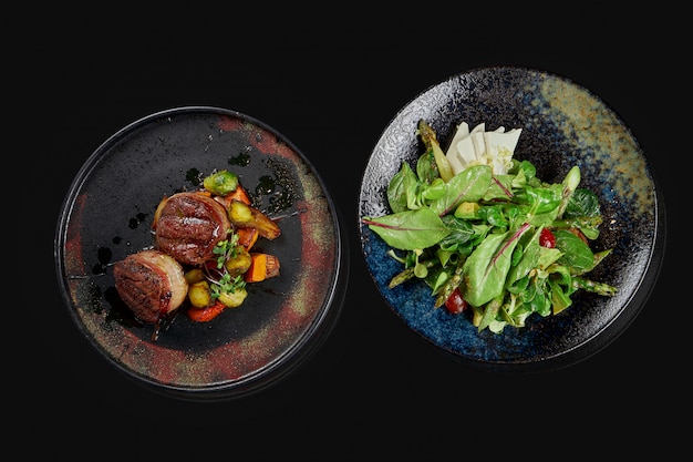 Foto dois pratos apetitosos - salada com queijo feta e tomate e bifes em elegantes placas de cerâmica em uma superfície preta. vista do topo. foto de comida para o menu