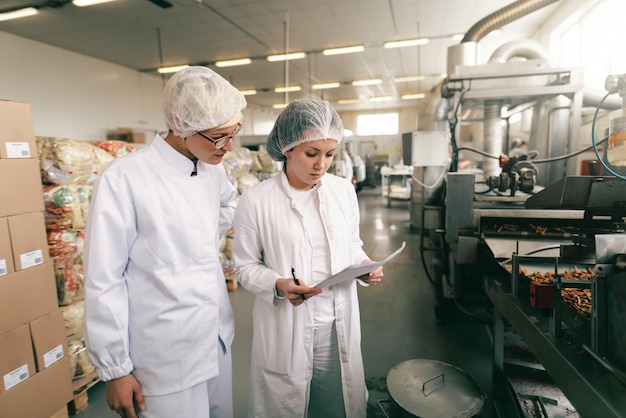 Dois profissionais de qualidade em uniformes estéreis brancos, verificando a qualidade de palitos de sal em pé na fábrica de alimentos.