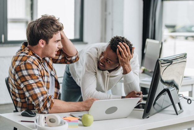 Foto dois programadores multiculturais chocados olhando um para o outro e segurando as mãos perto de cabeças