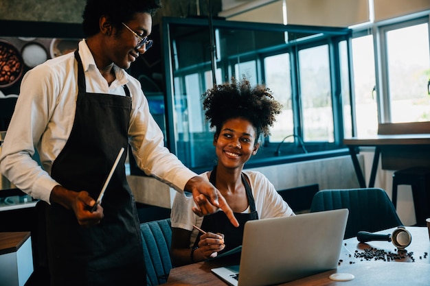 Dois proprietários de café barista preto e proprietário discutem o trabalho no laptop na cafeteria Planejamento de encomendas de suprimentos trabalho em equipe sucesso de negócios Cálculo de finanças discutindo o horário do menu