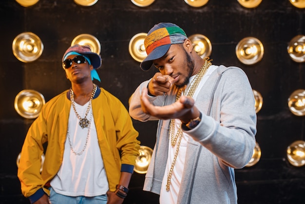 Dois rappers negros de boné, artistas no palco com holofotes