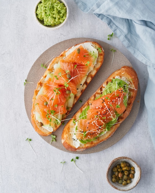 Dois sanduíches abertos, torradas com salmão, cream cheese, abacate, fatias de pepino no concreto branco