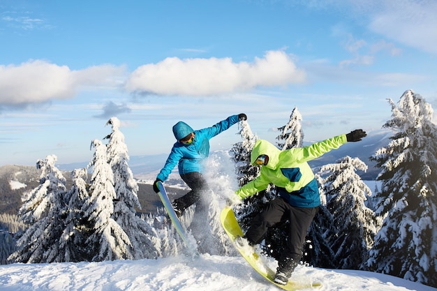 Dois snowboarders fazendo truques na estância de esqui Amigos de pilotos realizando salto com suas pranchas de snowboard perto da floresta na sessão de freeride sertão em roupa colorida e elegante Área de cópia