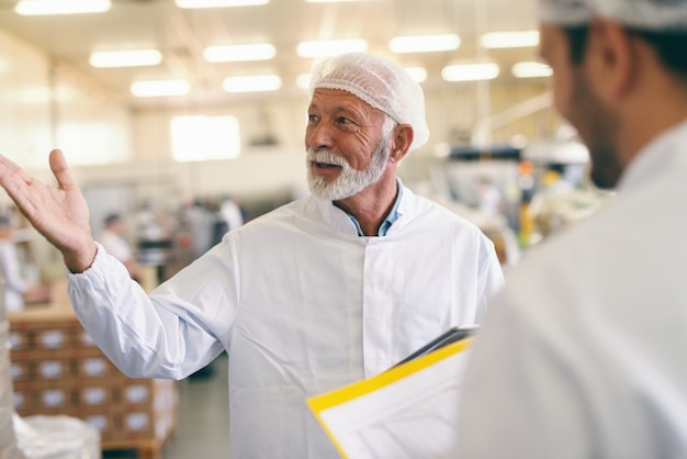 Dois trabalhadores caucasianos em traje de proteção, conversando e sorrindo em pé na fábrica de alimentos.