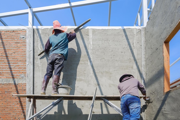 Dois trabalhadores de construção em andaimes de madeira estão encrustando uma parede de concreto no local de construção da casa