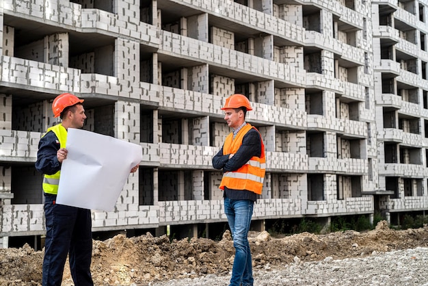 Dois trabalhadores fortes em uniformes de capacete com plano de construção em pé na construção de casas. Conceito de construção