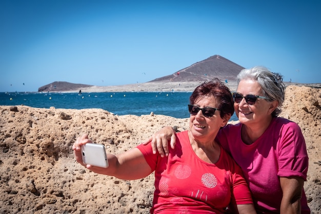 Dois velhos amigos sentados em um banco olham para o celular e tiram uma selfie. Conceito de vínculo e amizade. Montanha e mar atrás deles. Paraíso para os amantes do surf