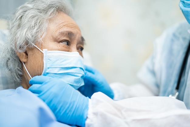 Doktor, der asiatischen älteren Fraupatienten prüft, der eine Gesichtsmaske trägt