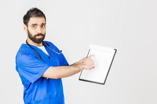Doktor im blauen Gewand mit dem Stethoskop, das auf Klemmbrett lokalisiert auf weißem Hintergrund zeigt