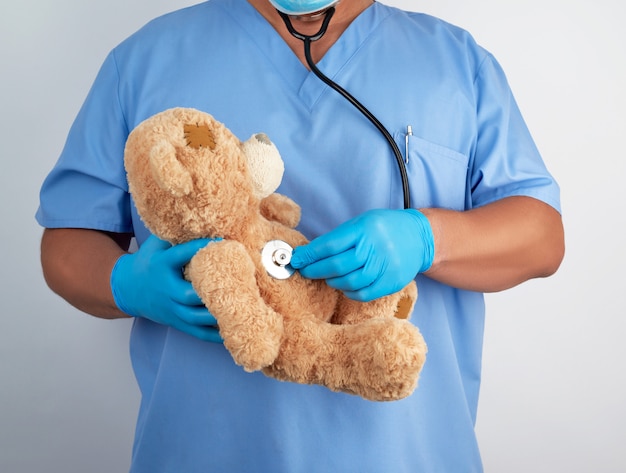 Doktor in blauer Uniform und weißen Latexhandschuhen hält einen braunen Teddybär,