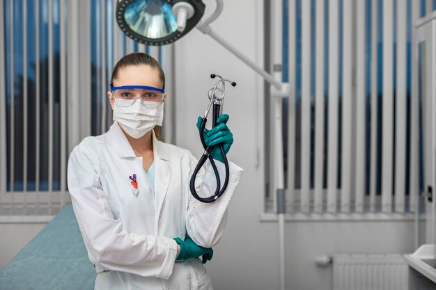 Doktor in einem weißen Kittel in einer Maske mit Brille und Handschuhen, die ein Phonendoskop vor dem Hintergrund eines Krankenzimmers halten.