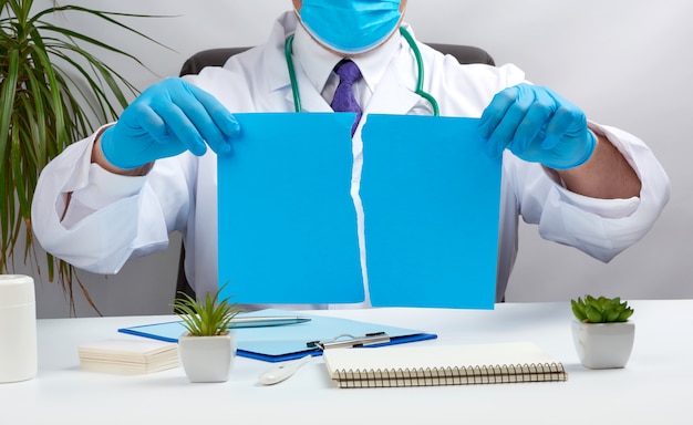 Doktor in einem weißen medizinischen Mantel sitzt an einem Tisch in einem braunen Ledersessel und hält ein zerrissenes blaues Blatt Papier