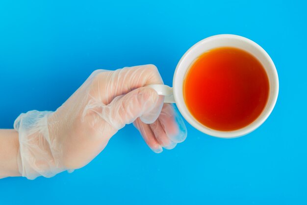 Doktorhand im medizinischen Handschuh hält weiße Teetasse auf dem blauen Hintergrund flatlay