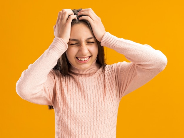 Dolor bastante adolescente manteniendo las manos en la cabeza con los ojos cerrados aislado en la pared naranja con espacio de copia