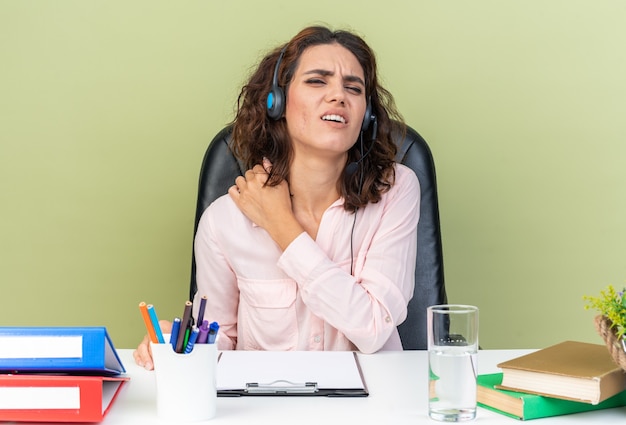 Dolor, bastante, caucásico, mujer, operador de call center, en, auriculares, sentado, en el escritorio, con, oficina, herramientas, poniendo, ella, hombro