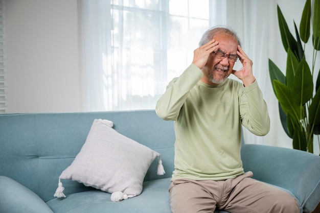 Dolor de cabeza hombre anciano asiático triste dolor de migraña crónica masajeando anciano sostiene la cabeza con la mano para el dolor de cabeza enfermedad en la sala de estar en casa problemas de salud de la vejez atención médica