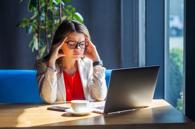 Dolor de cabeza, migraña, confusión, problema o pensamiento. Retrato de una joven morena enferma con gafas sentada, sosteniendo su dolorosa cabeza hacia abajo y sintiéndose mal. foto de estudio interior, cafetería, fondo de oficina