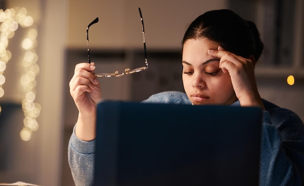 Dolor de cabeza de mujer negra y computadora portátil de oficina en casa de un estudiante con estrés y agotamiento Proyecto universitario en línea nocturno y ansiedad de una mujer joven con anteojos y fondo borroso en la oscuridad