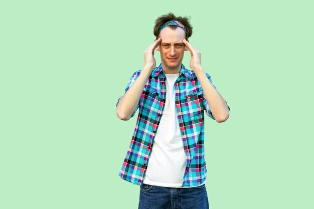 Dolor de cabeza o confundir. Retrato de un joven triste o enfermo con un pantalones a cuadros azul casual y una diadema de pie y sosteniendo su cabeza y sintiéndose mal. tiro de estudio interior, aislado sobre fondo verde claro