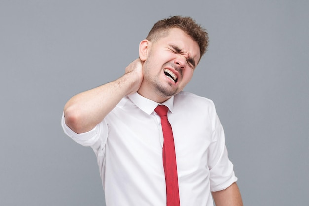 Dolor de cuello Retrato de un joven enfermo con camisa blanca y corbata de pie y tocando o masajeando su doloroso cuello interior aislado en fondo gris