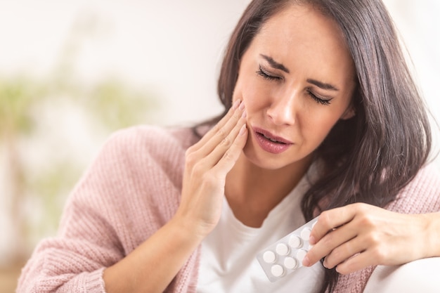 Dolor en los dientes mientras la morena sostiene su mejilla con una mano y pastillas analgésicas en la otra.