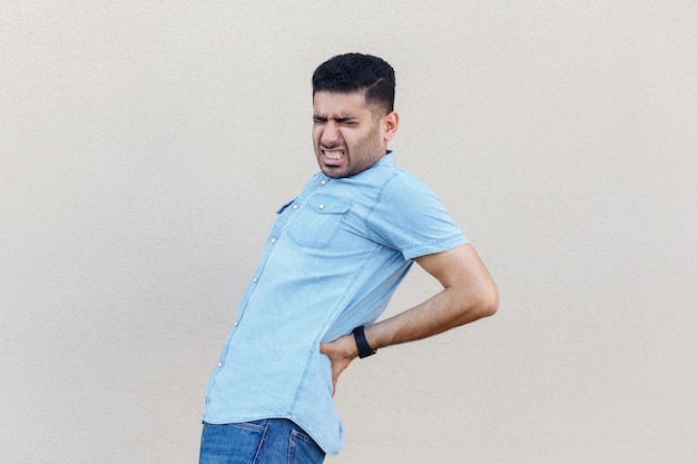 Dolor de espalda o dolor de riñón. Retrato de un joven apuesto y enfermo con barba y camisa azul de pie y sosteniendo su dolorosa espalda. tiro de estudio interior aislado sobre fondo de pared beige claro.