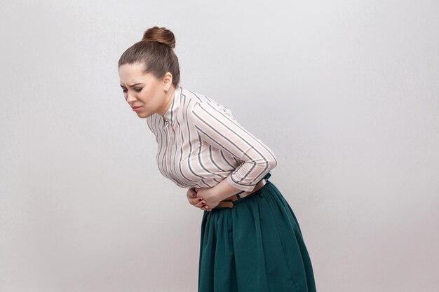 Dolor de estómago. Retrato de mujer joven hermosa en camisa a rayas y falda verde con peinado de prohibición recogido, de pie y sosteniendo su vientre doloroso. tiro del estudio de interior, aislado en fondo gris.