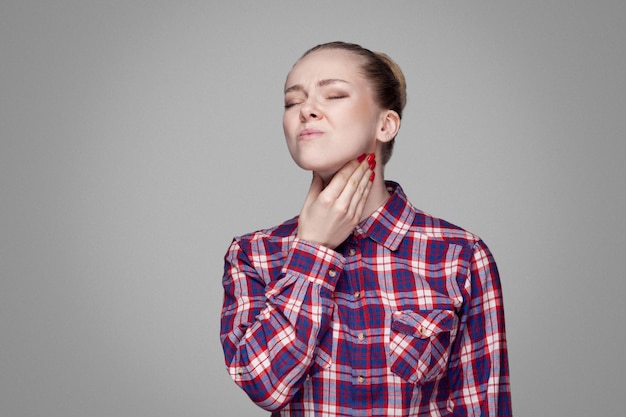 Foto dolor de garganta. chica rubia enferma con camisa a cuadros roja y rosa, peinado de moño recogido, maquillaje de pie y tocando su cuello doloroso. tiro del estudio de interior. aislado sobre fondo gris