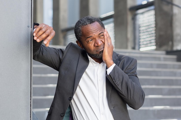 Dolor de muelas en la calle, un anciano afroamericano con traje se para fuera de una oficina y sostiene