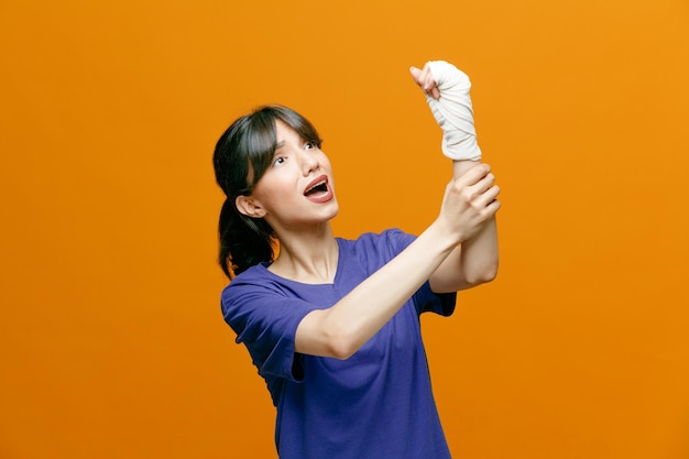 Dolorida jovem desportiva vestindo camiseta levantando a mão olhando para ela enquanto agarrava o braço com o pulso envolto com curativo isolado em fundo laranja